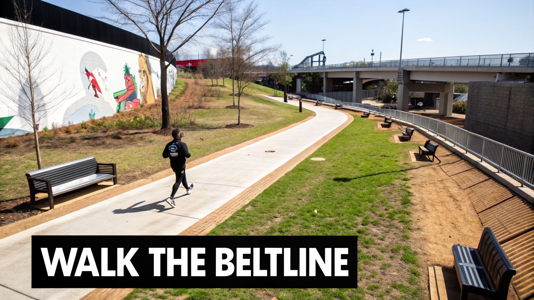 A person walks on the paved Atlanta Beltline trail, lined with grass, benches, and a colorful mural.