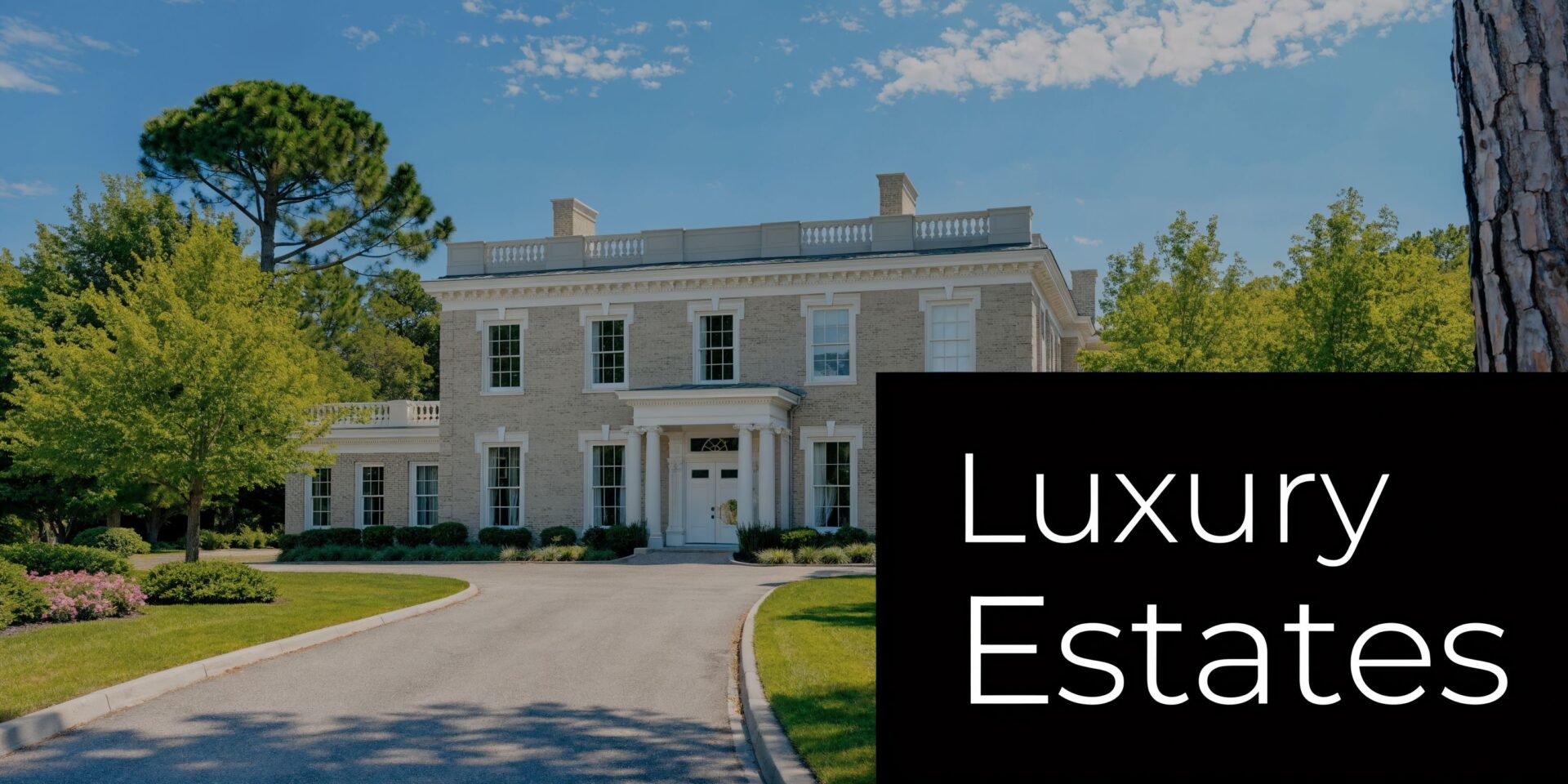 A beautiful classic brick mansion surrounded by lush green trees and a paved driveway under a blue sky.